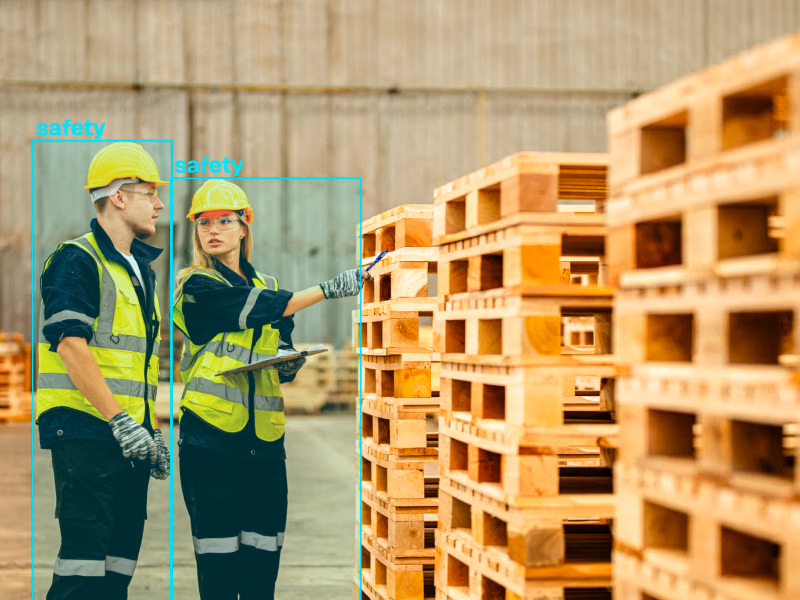 Two warehouse workers in yellow safety helmets and reflective vests detected with bounding boxes beside stacked wooden pallets.